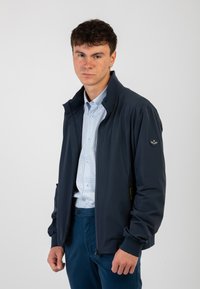 Giacca leggera blu navy con colletto rialzato, tasche con zip e polsini elasticizzati. Indossata sopra una camicia a righe azzurra.