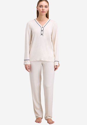 Woman standing barefoot wearing cream-colored long-sleeve top with black trim and matching pants against white background.
