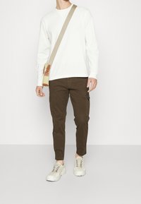 Bílá dlouhá rukávová tričko s crewneck výstřihem, hnědé cargo kalhoty se bočními kapsami a bílé tenisky se šedými akcenty. Béžová taška přes rameno s hnědými detaily.