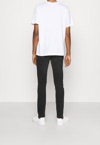 Man som står vänd mot väggen, iklädd en vit kortärmad t-shirt, svarta slim fit-jeans och vita sneakers på en heltäckande golv.