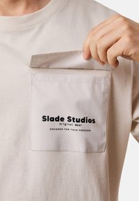 Beige t-shirt med en fyrkantig ficka, med svart text: "Slade Studios Original Wear" och "DESIGNED FOR YOUR FREEDOM." Slät tyg.