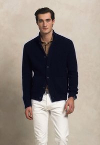 Jauns vīrietis, tērpies tumši zila pogājamā džemperī, virs brūnas krekla un baltām biksēm, stāv neitrālā fonā.