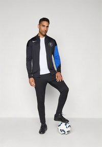 Schwarze Sportjacke mit einem blauen Ärmel, kombiniert mit schwarzen Hosen und schwarzen Fußballschuhen, steht auf einem weißen Fußball.