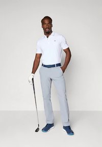 Balti polo marškinėliai, šviesiai pilkos kelnės, mėlynas diržas ir golfo batai. Laiko golfo lazdą, stovi prieš neutralią foną.