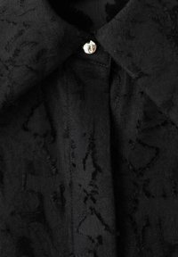 Camicia nera con texture e un motivo sottile, dotata di colletto a punta e un bottone argentato lucido all'apertura del collo.