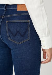 Donkerblauwe denim jeans hebben een kenmerkend "W"-ontwerp gestikt op de achterzak, met een tan leren label op de tailleband.