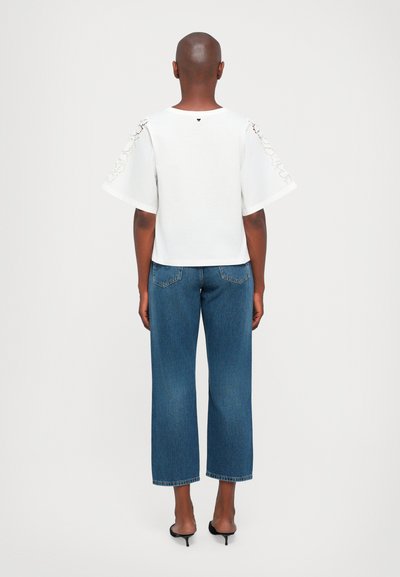 Valge topp laiade varrukatega, millel on pitsi detailid, koos tumeda pesu ja laia lõikega teksadega. Minimalistlik disain, ilma nähtavate logodeta.