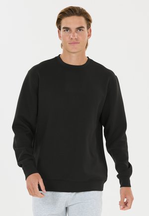 Junger Mann mit hellbraunen Haaren, der einen schlichten schwarzen Langarm-Sweatshirt und hellgraue Hose vor einem einfarbigen weißen Hintergrund trägt.
