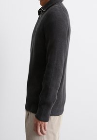 Dunkelgrauer, gerippter Strickpullover mit Reißverschluss am Kragen und langen Ärmeln, mit figurbetonter Passform und strukturierter Stoffqualität. Getragen über hellen Hosen.