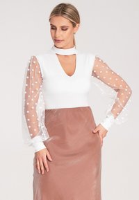 Biały top z głębokim dekoltem w kształcie litery V i przezroczystymi rękawami w kropki, zestawiony z satynową brązową spódnicą. Teksturowany materiał o miękkim układzie.