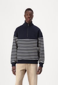 Navy and white striped zip-up sweatshirt with a high collar, featuring a relaxed fit and ribbed cuffs and hem. Worn with beige pants.