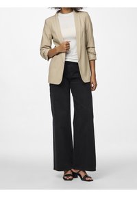Blazer beige avec col châle, associé à un t-shirt blanc et un jean noir taille haute à jambes larges. Les chaussures sont des sandales à talons noires.