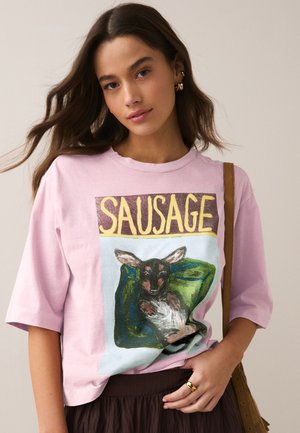 Kvinde med langt brunt hår iført en pink T-shirt med en tegning af en sort hund, der ligger på en grøn pude, og ordet "SAUSAGE" ovenover.