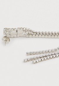 Brincos de metal prateado em forma de studs com ligações de corrente em forma de cubo penduradas em três fios, sobre um fundo branco.