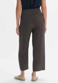 Kobieta w wysokich, krótkich spodniach z geometrycznym wzorem w diamenty, w połączeniu z beżowymi płaskimi butami i czarną bluzką, przedstawiona od tyłu.