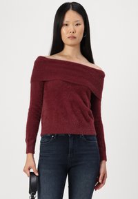 Pulover burgundy cu umeri descoperiți, cu mâneci lungi, textură moale și un decolteu în straturi, asortat cu blugi skinny negri.