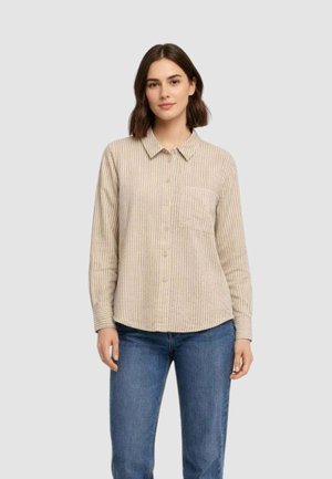 Donna che indossa una camicia a righe verticali beige e bianche con taschino frontale e jeans blu su sfondo uniforme.