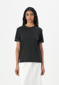 T-shirt noir en coton à manches courtes avec un col rond, présentant des coutures contrastées sur les bords. Associé à une jupe blanche à motifs.