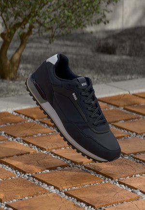 Czarny sneaker BOSS z białą podeszwą i sznurówkami, spoczywający na drewnianych kwadratowych płytkach na zewnątrz, blisko drzewa i żwiru.