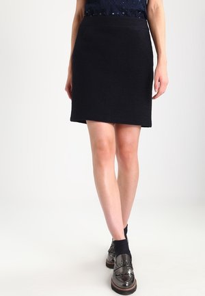 Mini-jupe noire, texturée, avec une coupe droite et une taille, stylée avec des mocassins noirs brillants et des chaussettes noires jusqu'à la cheville.