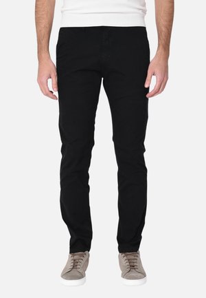 Uomo con pantaloni neri aderenti e sneaker grigio chiaro, in piedi davanti a uno sfondo bianco neutro.