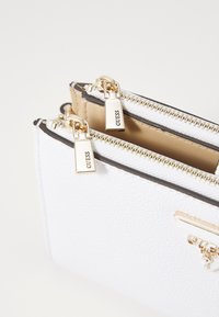 Valge tekstuuriga rahakott kuldse tõmblukuga, millel on kaks sektsiooni ja kolmnurkne kuldne aksessuaar brändi nimega "Guess."