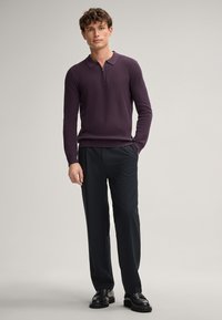 Lila Sweatshirt mit Reißverschlusskragen aus strukturiertem Strick, kombiniert mit schwarzen Hosen. Das Model trägt schwarze Schuhe und steht vor einem neutralen Hintergrund.