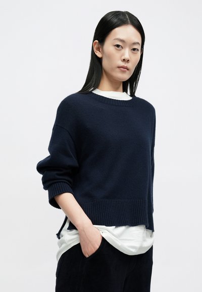 Πλεκτό πουλόβερ σε ναυτικό μπλε με στρογγυλή λαιμόκοψη, άνετη εφαρμογή, ριμπ τελείωμα και χαμηλωμένους ώμους, φορεμένο πάνω από ένα λευκό πουκάμισο.