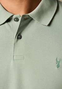 Polo de color verde claro hecho de tela texturizada, que presenta un collar doblado, dos botones y un pequeño logotipo de ciervo bordado en el pecho.