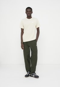 Μπεζ βαμβακερό t-shirt με μικρό λευκό λογότυπο, συνδυασμένο με χακί cargo παντελόνι με πλαϊνές τσέπες και μαύρα αθλητικά παπούτσια με λευκές λεπτομέρειες.