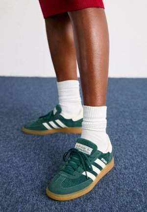 Des jambes portant des chaussettes blanches et des baskets vertes Adidas Handball Spezial sur un tapis bleu.