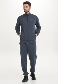 Dunkelgrauer Trainingsanzug mit hohem Kragen, Frontreißverschluss und zwei Taschen. Der Stoff wirkt weich mit einer subtilen Textur. Schwarze Sportschuhe werden getragen.