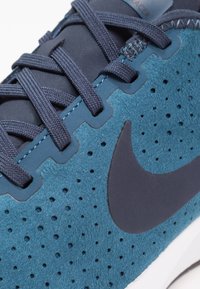 Tyrkysová sportovní obuv s perforovaným semišovým svrškem, tmavě modrými tkaničkami a černým logem. Má polstrovanou bílou podrážku a textilní detaily.