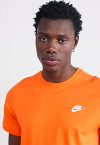 Oranžs Nike T-krekls ar apaļu kaklu, izgatavots no kokvilnas, ar baltu logo kreisajā krūšu pusē. Īsas piedurknes un gluda tekstūra.