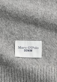 Grijs gebreide stof met een vlakgebreide textuur, voorzien van een wit label met blauwe tekst: "Marc O'Polo DENIM." Close-up detail van de textuur.