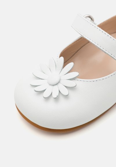 Zapato Mary Jane de cuero blanco con un adorno de flor de margarita, puntera redondeada y suela de goma color marrón. Correa ajustable con hebilla plateada.