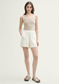 Beige linne med rundad halsringning, kombinerat med vita shorts med svarta och bruna vertikala ränder. Skorna är i form av giltiga sandaler.