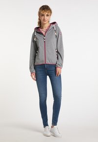 Giacca grigia con zip, dettagli rosa, cappuccio con cordino e tasche laterali. Indossata con jeans skinny blu e sneakers bianche. Camicia a righe sotto.
