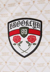 Nášivka s designem štítu, na kterém je napsáno "BROOKLYN" černou barvou, červený pruh, zelené pozadí a dvě červené růže na stoncích, zasazené do vzorovaného krémového pozadí.
