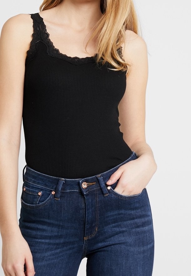 Frau in einem schwarzen, gerippten Tanktop mit Spitzenbesatz und hochgeschnittenen dunkelblauen Jeans, mit der Hand in der Tasche, vor einem schlichten Hintergrund.