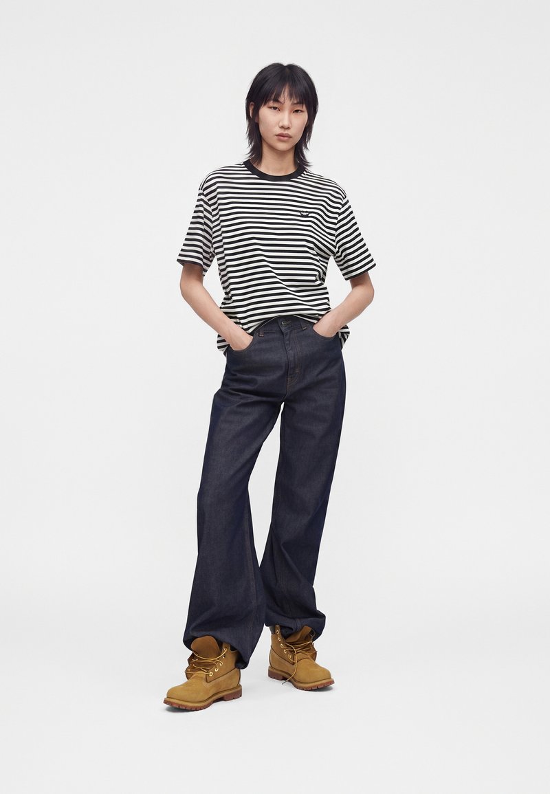 Giovane donna in piedi con le mani nelle tasche, indossa una maglietta a righe bianca e nera, jeans larghi scuri e stivali da lavoro color cammello su sfondo bianco.