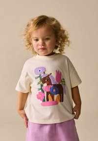 Krémové bavlněné tričko s barevným koněm a květinami v textilní aplikaci. Kombinováno s světle fialovými šortkami.