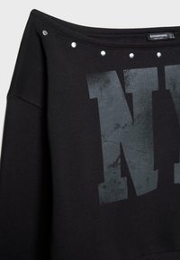 Crna majica s velikim izblijedjelim sivim slovom "N" na prsima, srebrnim okruglim zakovicama duž širokog izreza i dugim rukavima.