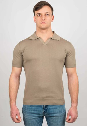 Giovane uomo fermo in piedi che indossa una polo beige aderente a maniche corte e jeans blu su uno sfondo chiaro e uniforme.