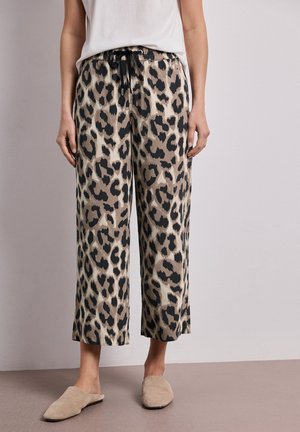 Persona que lleva pantalones anchos con estampado de leopardo en beige y negro, top blanco y zapatos beige tipo slip-on, de pie frente a una pared lisa.