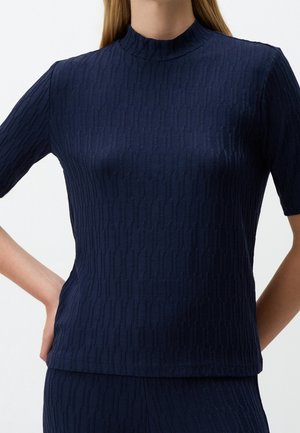 Vrouw draagt een marineblauw geribd gebreid topje met korte mouwen en een opstaande kraag, staand met één hand op haar heup.