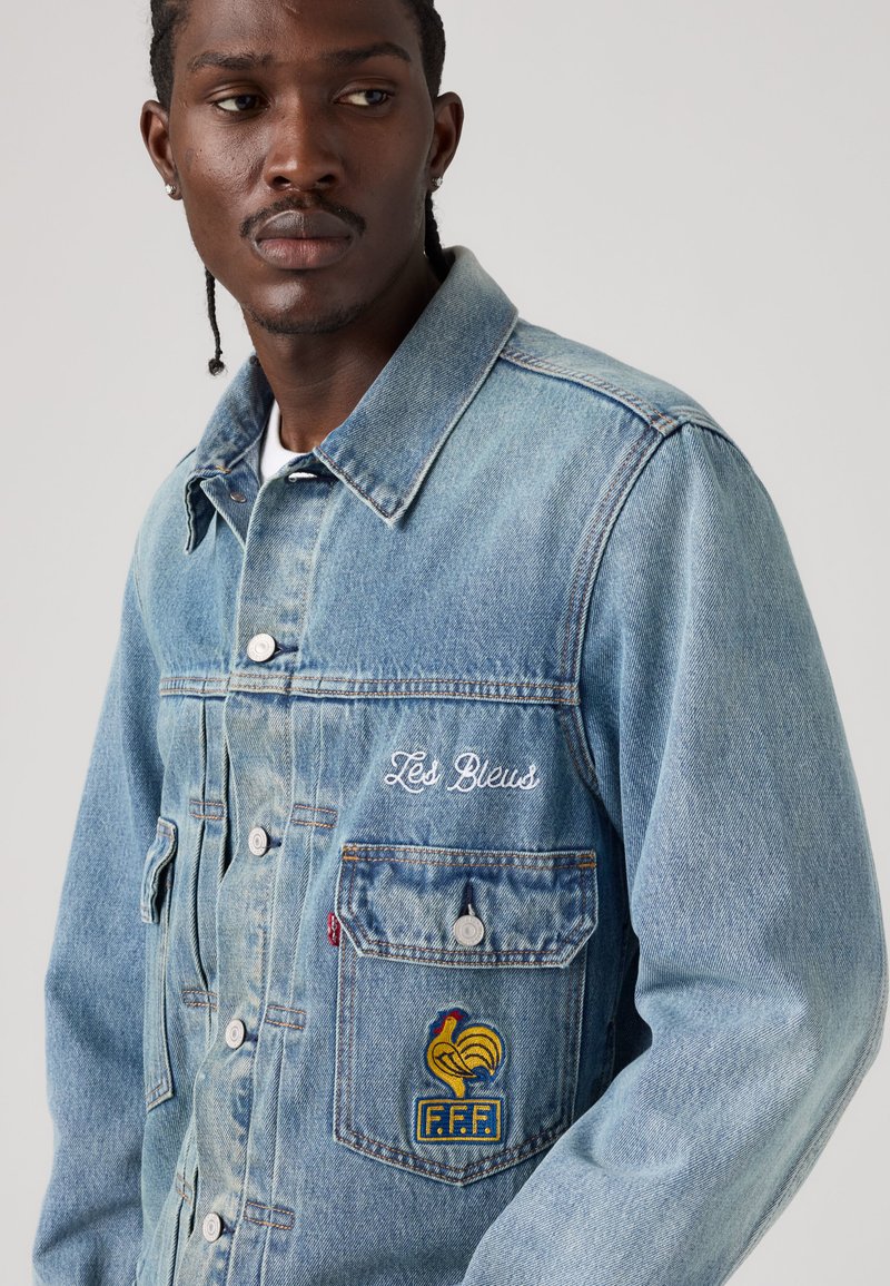 Uomo con capelli intrecciati che indossa una giacca di jeans azzurro chiaro con il testo ricamato "Les Bleus" e una toppa con il gallo F.F.F. sulla tasca.