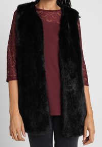 Schwarze Fleece-Weste mit offenem Frontbereich, weicher Textur und ohne Verschlüsse. Getragen über einem bordeauxfarbenen Oberteil mit Spitzendetails und Dreiviertelärmeln.