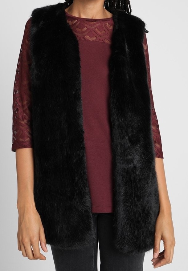 Schwarze Fleece-Weste mit offenem Frontbereich, weicher Textur und ohne Verschlüsse. Getragen über einem bordeauxfarbenen Oberteil mit Spitzendetails und Dreiviertelärmeln.