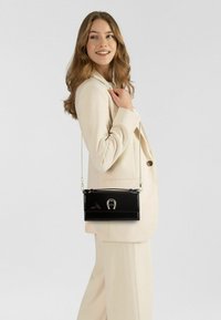 Bolso de mano de charol negro con cierre plateado, forma rectangular y correa de cadena desmontable, exhibido sobre un fondo claro.